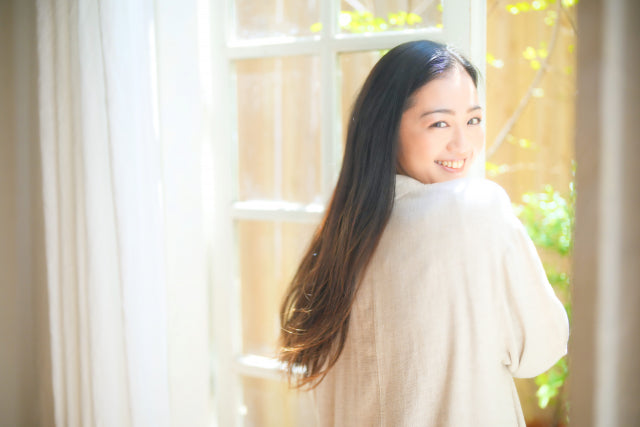 振り返りの笑顔が素敵な愛され肌の女性。窓枠のそばで白く薄い衣装を着ている。