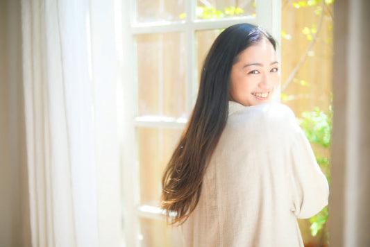 振り返りの笑顔が素敵な愛され肌の女性。窓枠のそばで白く薄い衣装を着ている。