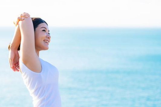 海辺で白いノースリーブ姿の肌の美しい女性が腕を曲げてストレッチしている